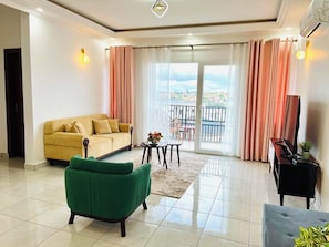 A living room with a sofa, a green armchair, a piano, and a balcony with a view. - 3K Residences Yaoundé Odza - Premium 2-Bedroom Apartment - D3 (Yaoundé)