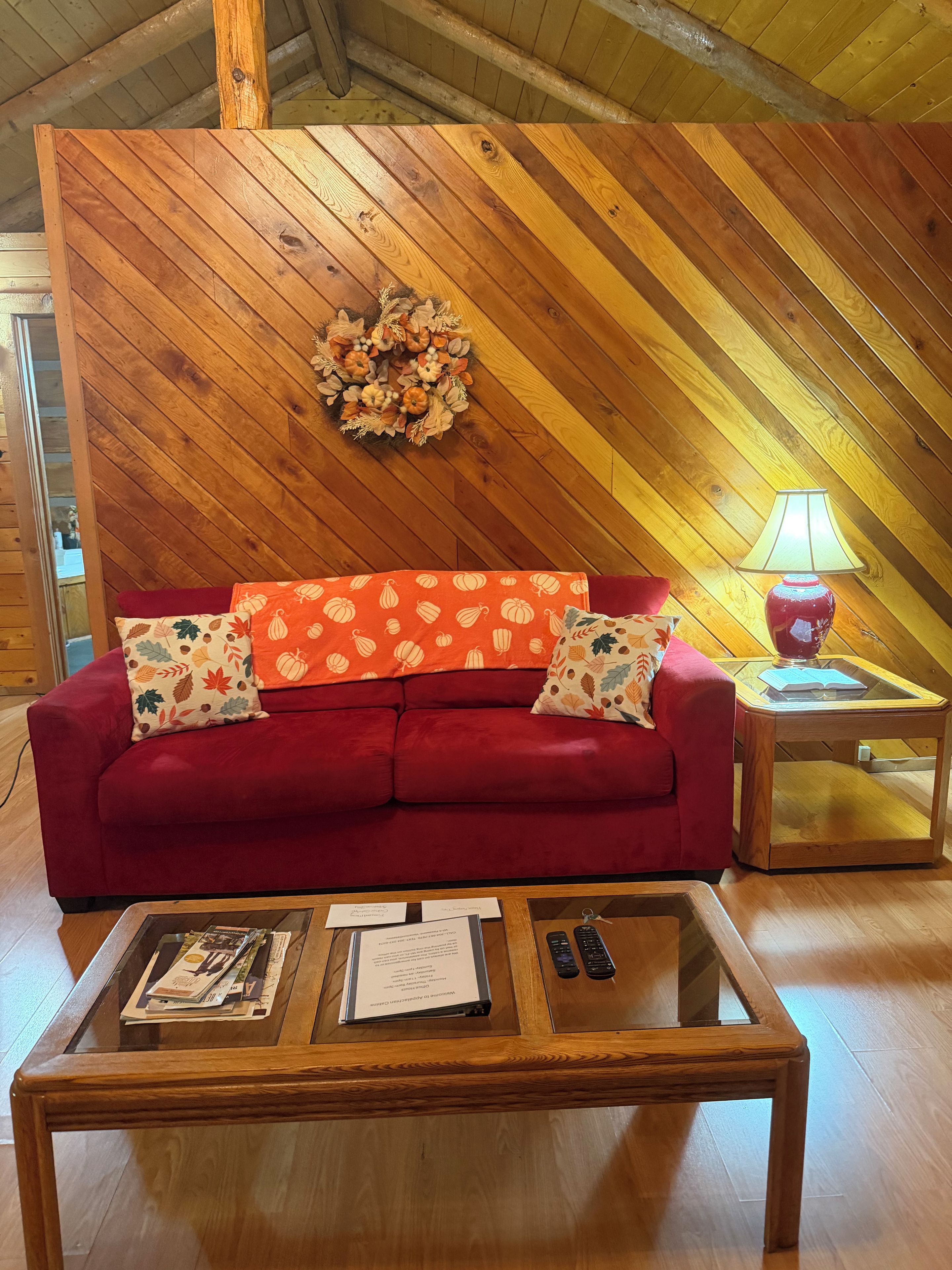 A red sofa with patterned cushions, a glass-top coffee table with magazines and a menu, and a wooden wall with a decorative wreath.