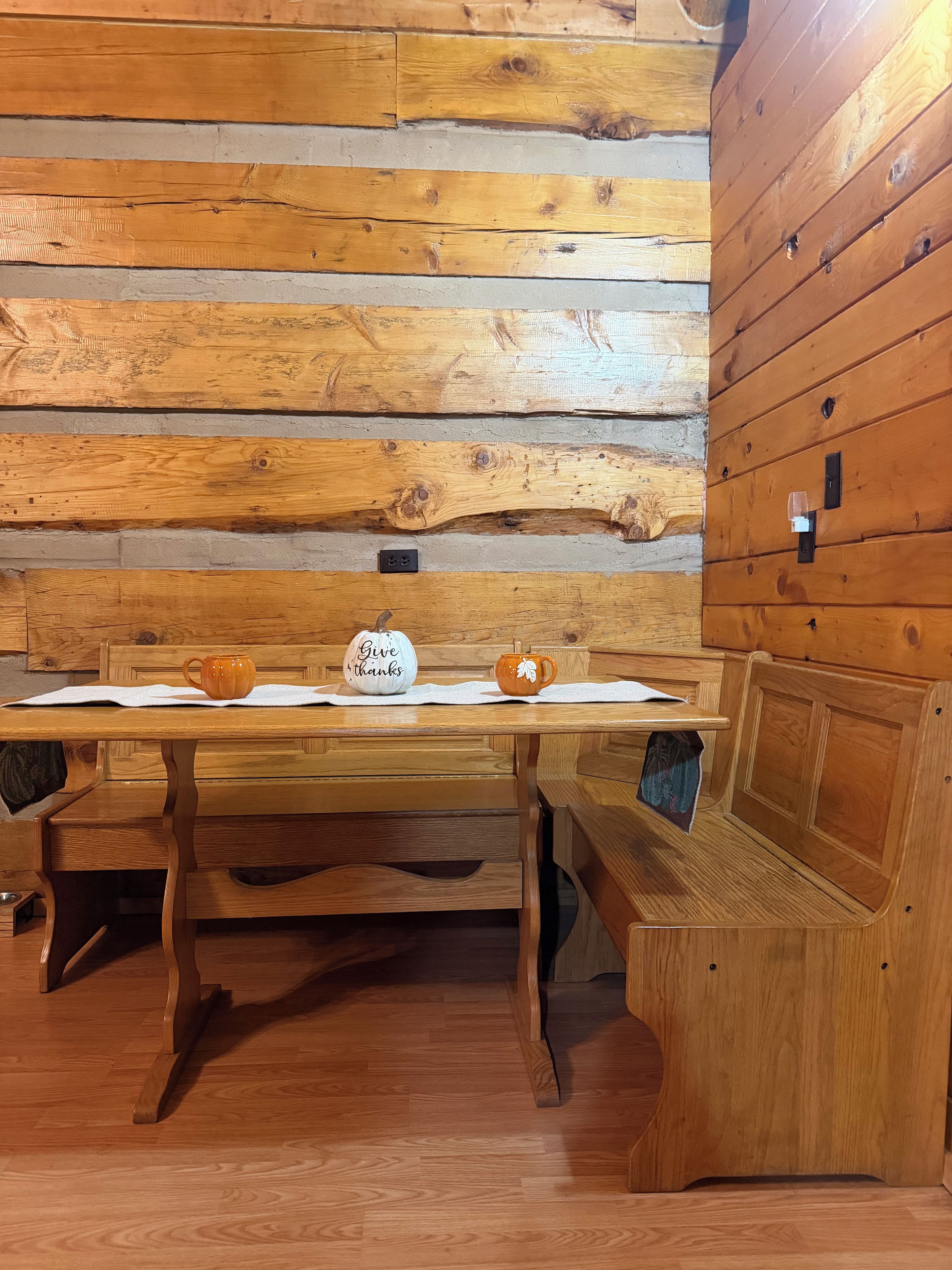 A wooden table with two mugs and a sign that says 'Give thanks' in a cabin-style room with wooden walls and benches.