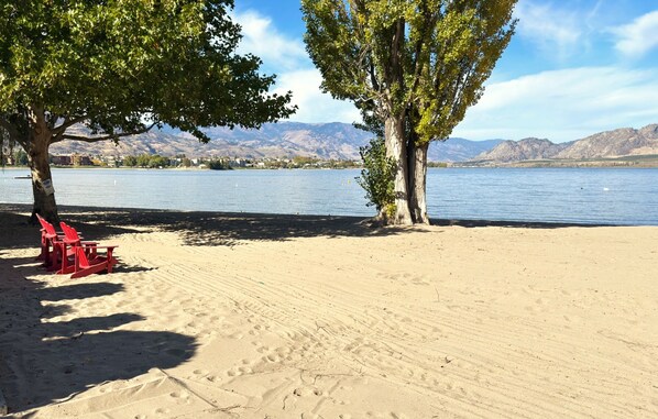 Ein Sandstrand mit roten Liegestühlen und Bäumen, ein See sowie Berge im Hintergrund.