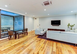 Una sala de estar moderna con piso de madera, una pared divisoria de vidrio, un sofá, un televisor y un comedor con sillas y una mesa.