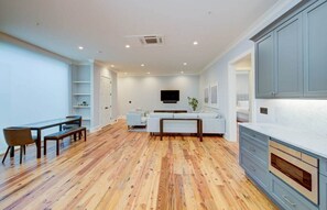 A spacious living area with a wooden floor, a sofa, a TV mounted on the wall, and a kitchen with grey cabinets. - Deluxe Two Bedroom Suite - Steps to King Street (Charleston)