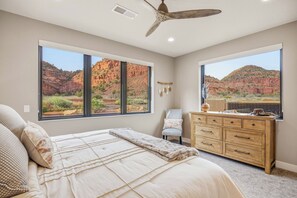 A bedroom with a bed, a dresser, and two windows offering a view of a desert landscape.