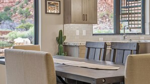 A dining area with a wooden table and chairs, a kitchen with cabinets and a window overlooking a garden. - Grand Kanab'n: Rooftop Hot Tub, Red Rock Views (Kanab)
