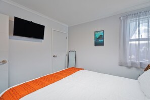 A bedroom with a bed, a television mounted on the wall, a door, and a window with curtains. - Endeavour Oasis - Whitianga Holiday Home (Whitianga)