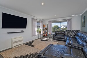 A modern living room with a black leather sofa, a flat-screen TV mounted on the wall, and a large window with curtains. - Endeavour Oasis - Whitianga Holiday Home (Whitianga)