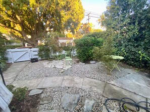 A backyard with a patio area, a white fence, and a large tree. - Charming Victorian Style 3-bedroom house with AC (Santa Monica)
