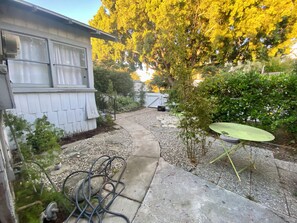 Un jardin à l’arrière d’une maison, avec un chemin de gravier, une petite table verte et une maison blanche dotée d’une fenêtre.