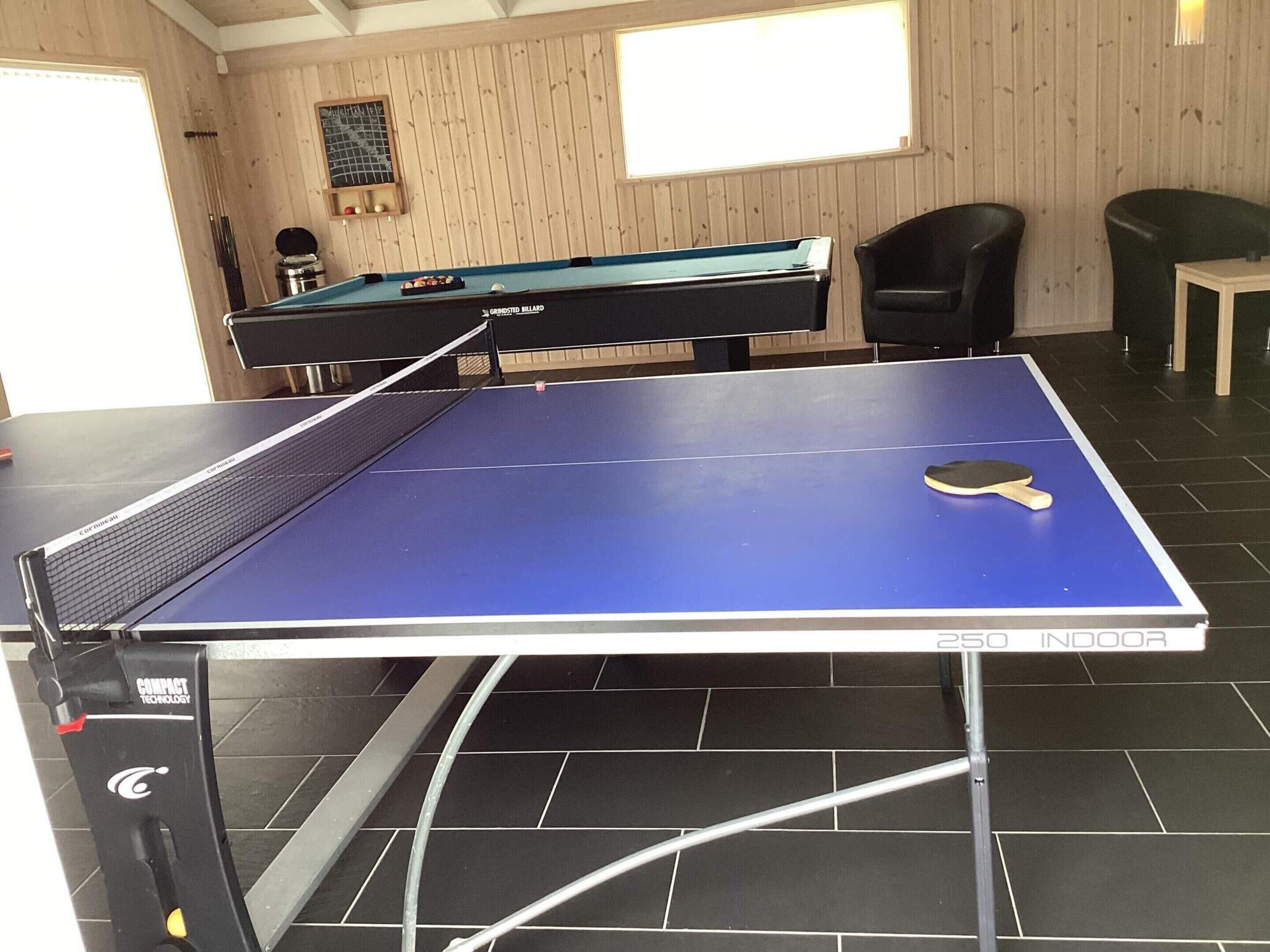 Une table de ping-pong avec un filet, une raquette de tennis de table, et une table de billard en arrière-plan.