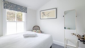 A bedroom with a bed, a window with curtains, a framed picture on the wall, a mirror, and a small plant. - Stunning 3Bed Victorian Central Home (Cambridge)