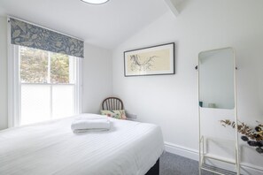Ein Schlafzimmer mit Bett, einem Fenster mit Vorhängen, einem gerahmten Bild an der Wand, einem Spiegel und einer kleinen Pflanze.