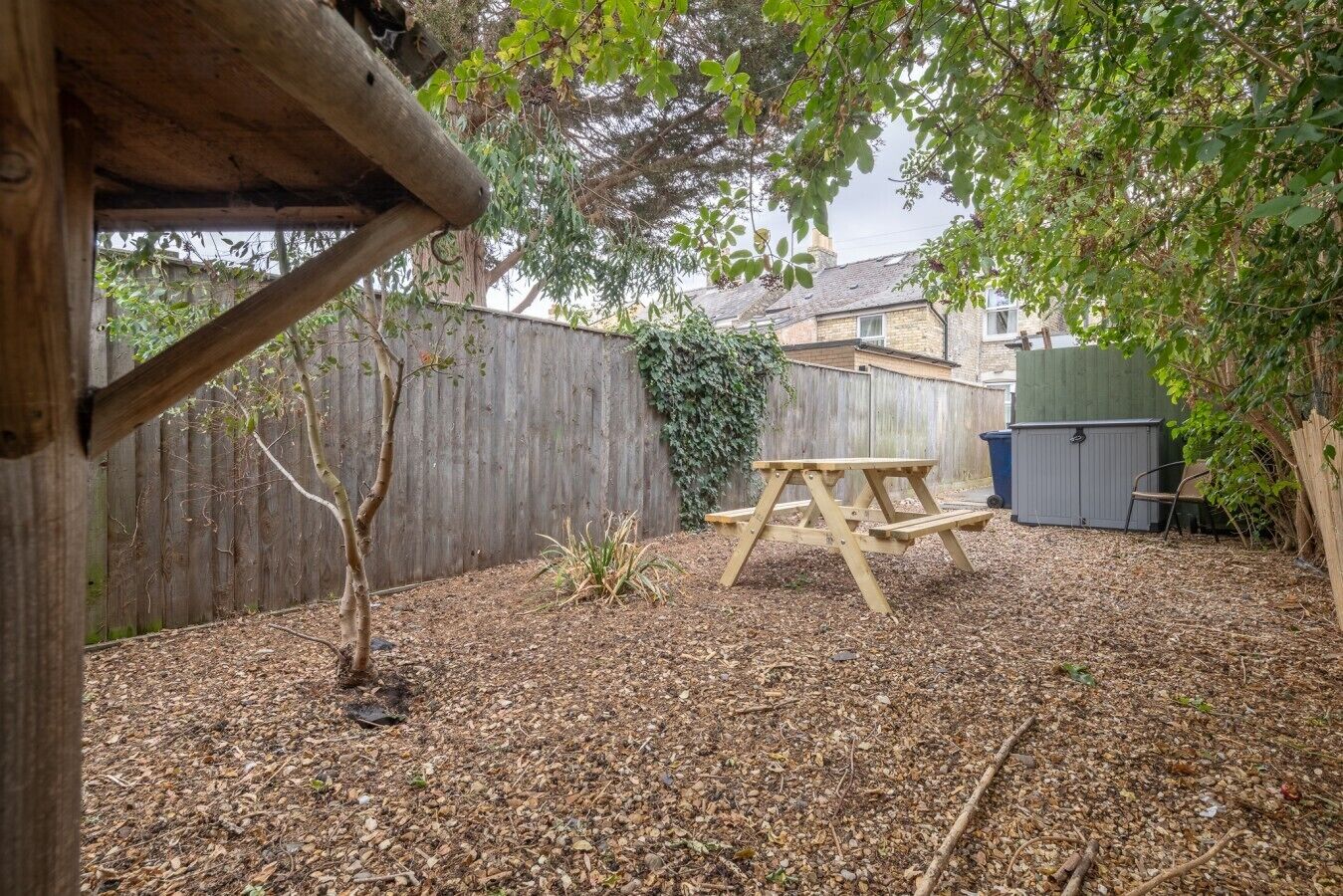 A wooden picnic table in a backyard with a wooden fence, a shed, and a tree.