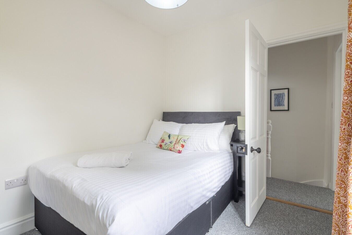 A neatly made bed with white linens and a gray headboard, a bedside table with a lamp, and a door leading to another room.