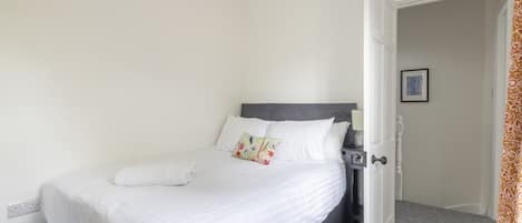 A neatly made bed with white linens and a gray headboard, a bedside table with a lamp, and a door leading to another room.