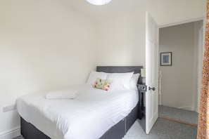 A neatly made bed with white linens and a gray headboard, a bedside table with a lamp, and a door leading to another room. - Charming 3Bed Victorian Home PLUS 1 Bed Annexe (Cambridge)