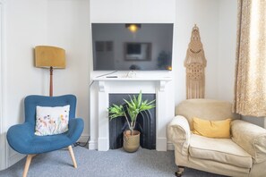 A living room with a television, a blue armchair, a beige sofa, a fireplace, and a decorative wall piece. - Charming 3Bed Victorian Home PLUS 1 Bed Annexe (Cambridge)