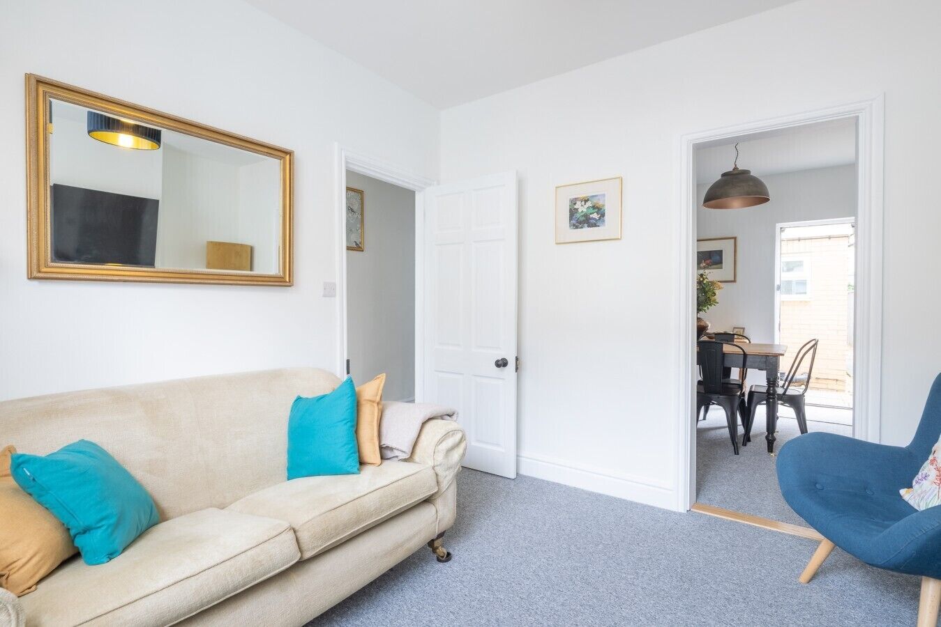 A living room with a beige sofa, a blue armchair, a dining area with a table and chairs, and a door leading to another room.