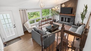 A modern living room with a fireplace, comfortable seating, and a large window offering a view of greenery. - Forest Escape Near Tremblant | Jacuzzi & Arcade (Mont-Tremblant)