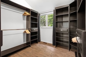 A walk-in closet with multiple shelves and drawers, a wooden hanger rack, and a window with a view of greenery.