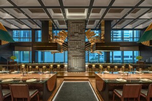 Un restaurante moderno con una columna central, varias mesas de comedor y grandes ventanales con vistas a la ciudad.