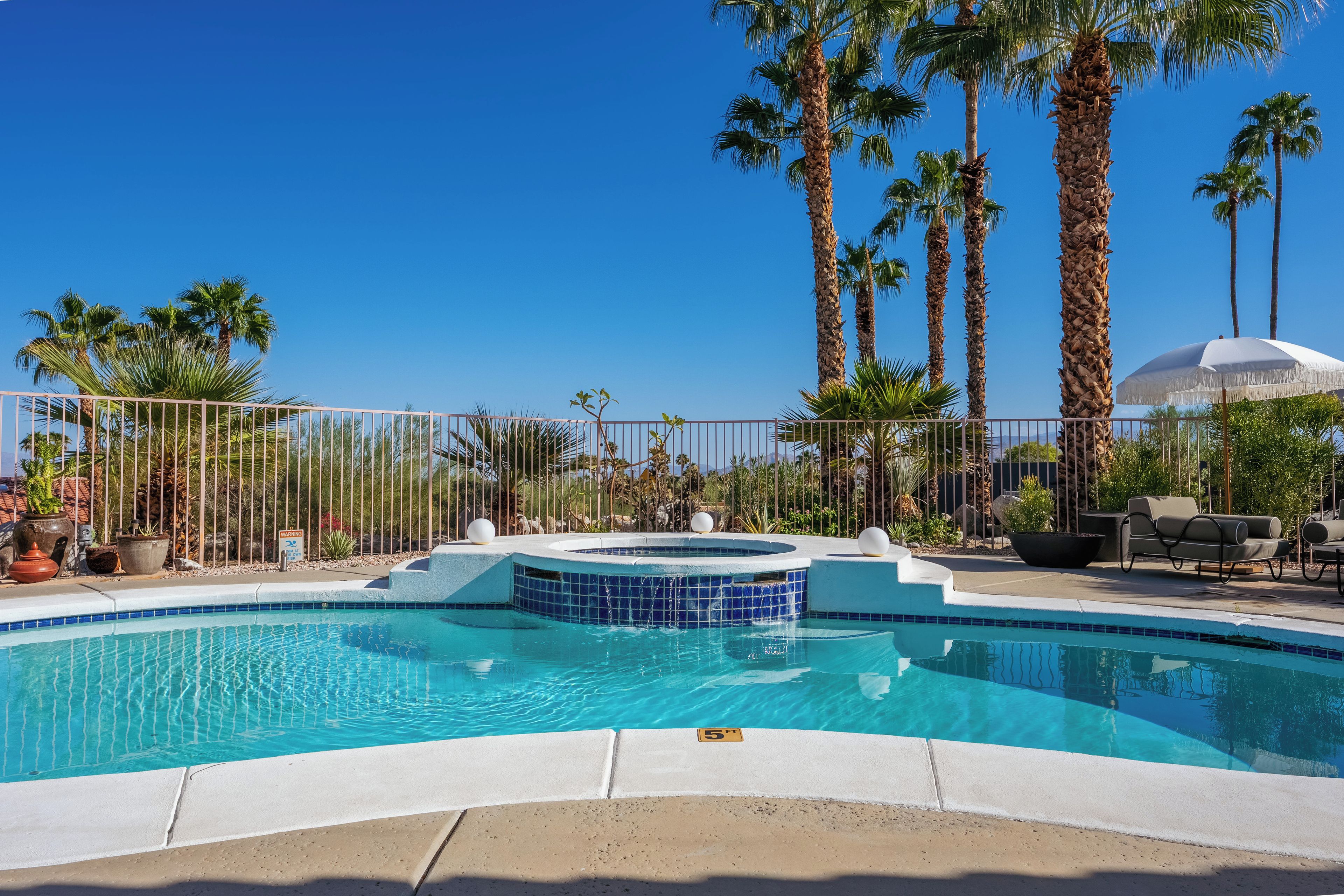 Une piscine avec un jacuzzi, entourée de palmiers et d’un espace clos aménagé avec des chaises longues.