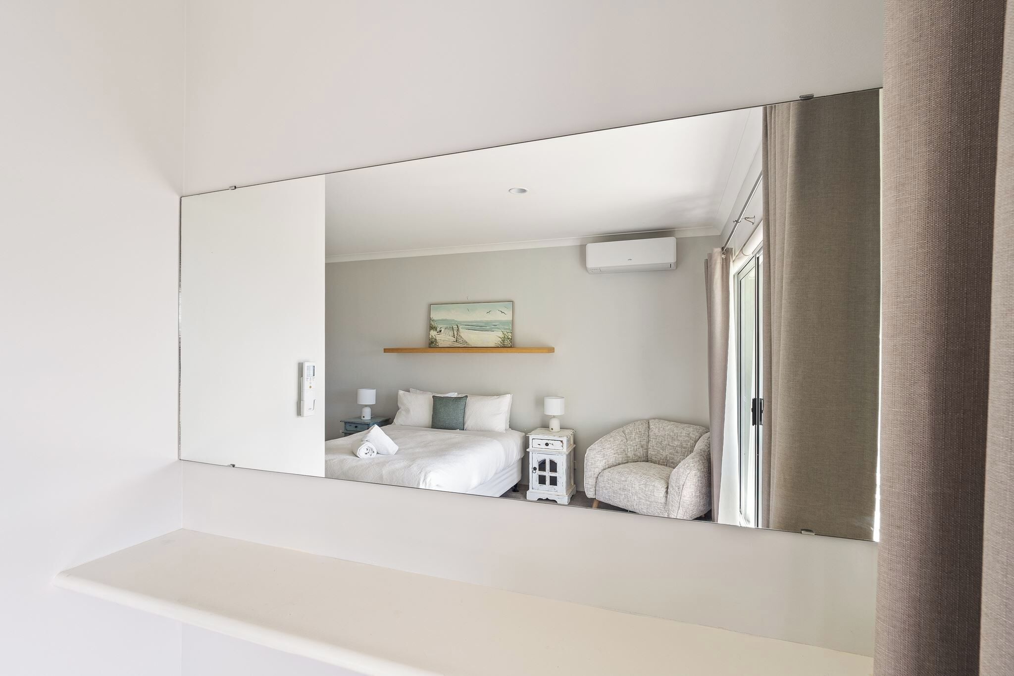 A bedroom with a large mirror, a bed, a chair, a bedside table, and a wall-mounted air conditioner.