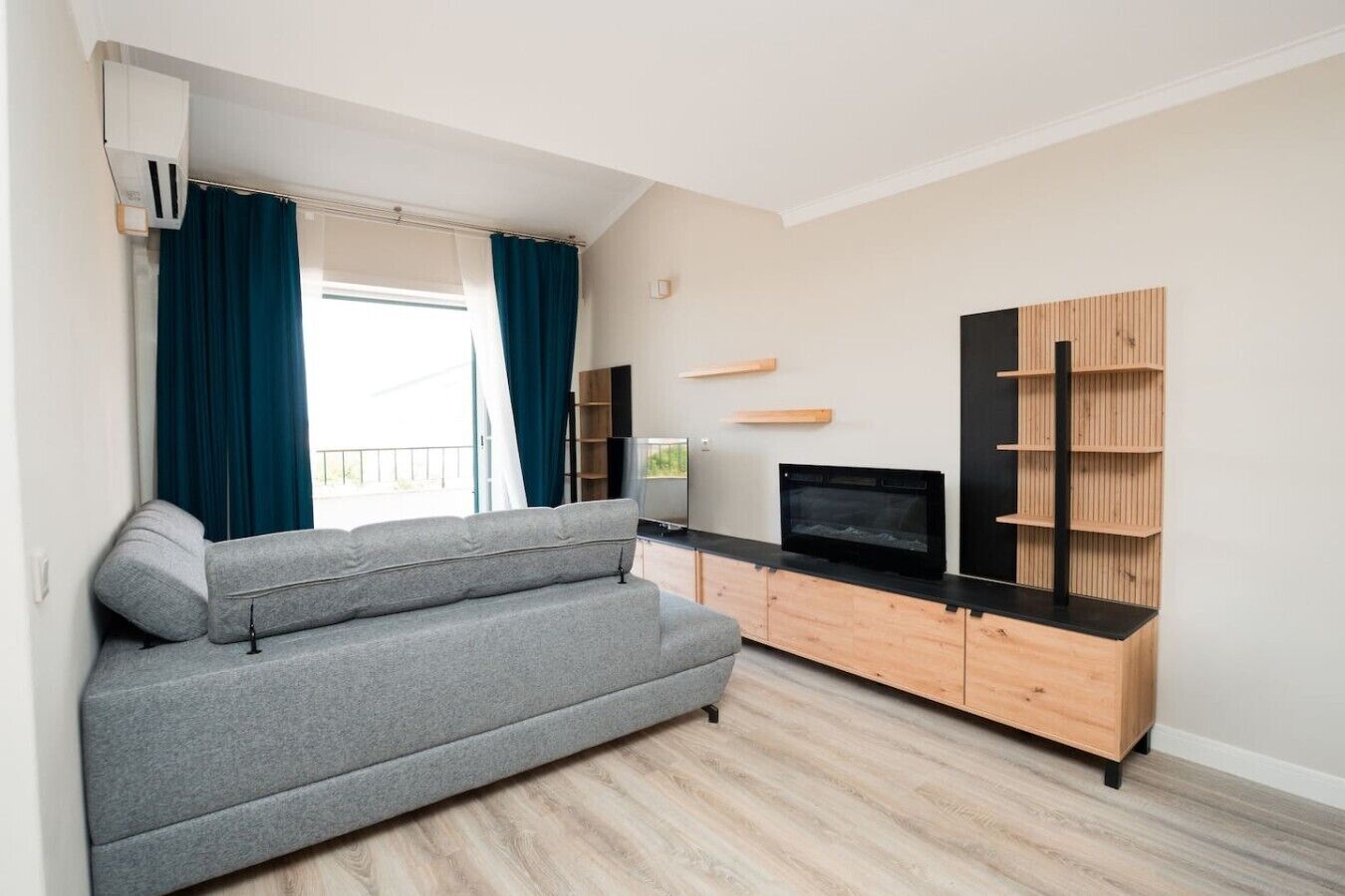 A modern living room with a grey sofa, a flat-screen TV, wooden shelves, and a window with curtains.