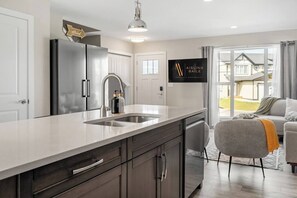 A modern kitchen with a stainless steel refrigerator, a built-in sink, and a dining area with a grey sofa and a view of the outside.