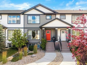 A two-story residential building with a red door, a front porch, and a small garden area.