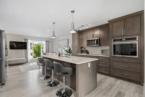 A modern kitchen with a central island, bar stools, stainless steel appliances, and a view of greenery outside.
