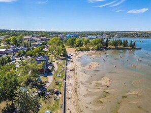 An aerial view of a beach with clear waters, a parking area, and a building with a sloped roof. - Red Door Retreat | Lake | Hot Tub | Fire Pit | AC (Sylvan Lake)