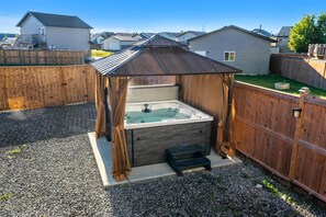 Un bain à remous doté d’une structure en bois, entouré d’une clôture en bois, d’un sol en gravier et de maisons en arrière-plan.