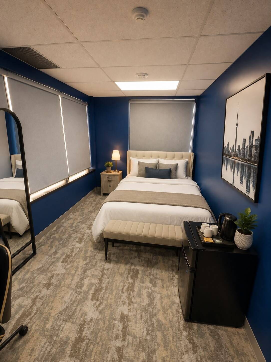 Quarto de hotel com uma cama grande, mesas de cabeceira, uma escrivaninha com cadeira, um espelho e uma pintura de paisagem urbana na parede.