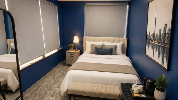 A hotel room with a large bed, bedside tables, a desk with a chair, a mirror, and a cityscape painting on the wall.