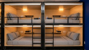 A bunk bed room with two levels, each with a mattress and pillows, and a shelf with a water bottle.