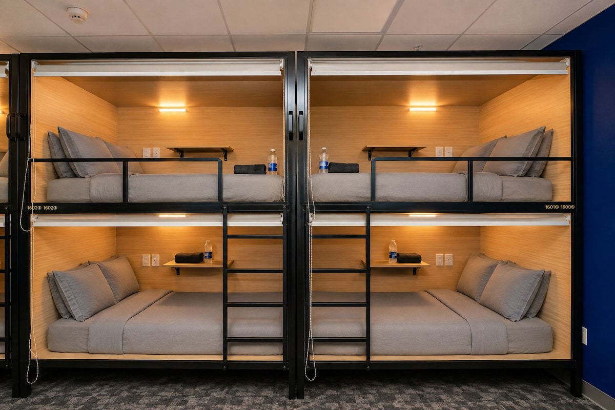 A bunk bed room with two levels, each with a mattress and pillows, and a shelf with a water bottle.