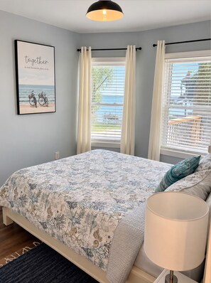 A bedroom with a bed, a bedside lamp, a window with curtains, a framed picture on the wall, and a view of the outdoors. - Crystal Beach Long Term Coastal Cottage (Fort Erie)