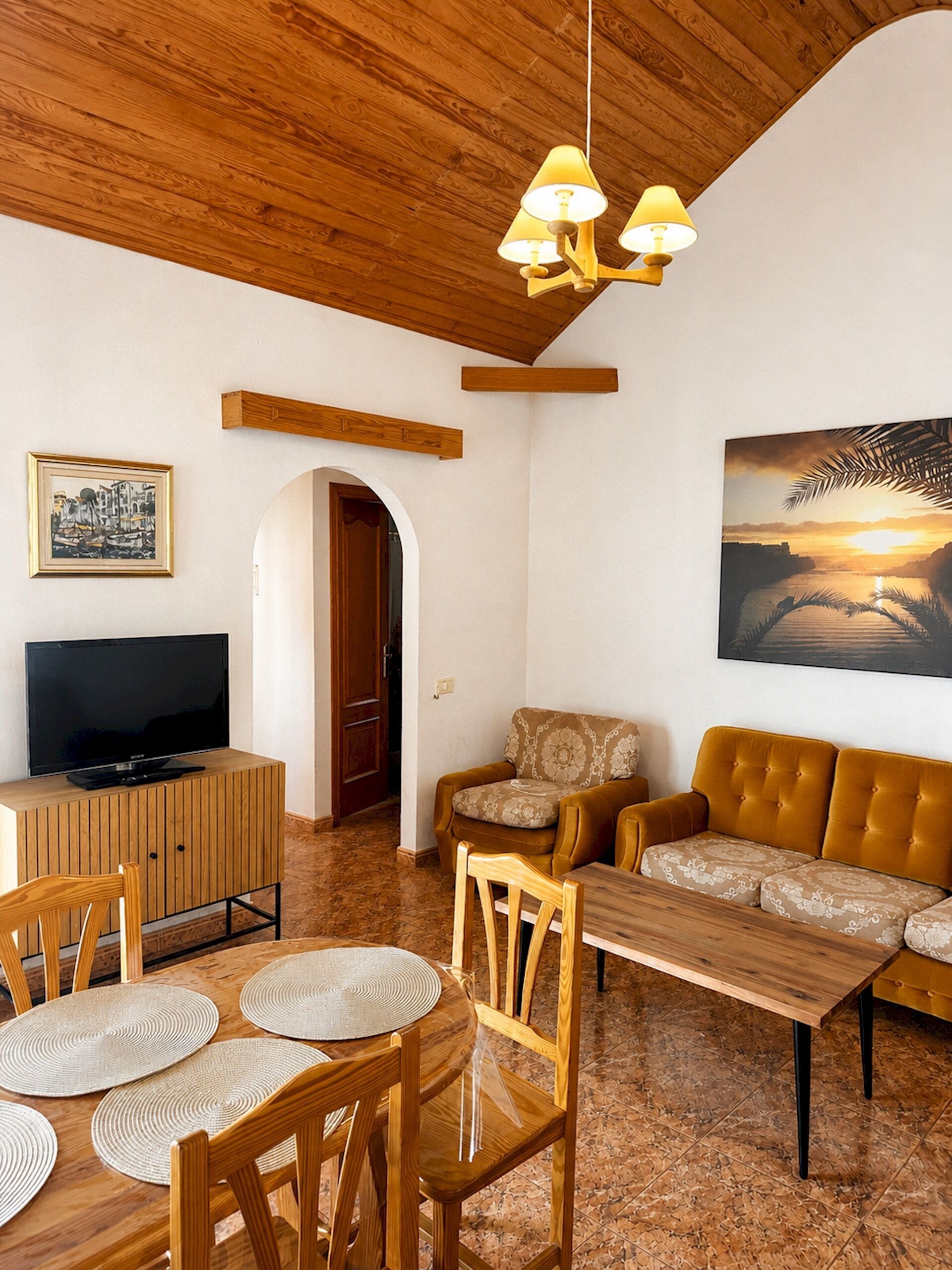 Un soggiorno con soffitto in legno, una televisione su un mobiletto in legno, un divano, un tavolo da pranzo con sedie e un quadro appeso al muro.