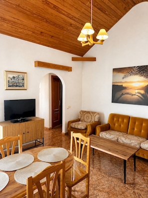 Ein Wohnzimmer mit Holzboden, einem Fernseher auf einer Holzkommode, einem Sofa, einem Esstisch mit Stühlen und einem Bild an der Wand.
