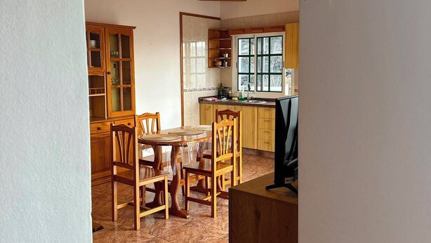 Ein Essbereich mit Holzkstühlen und einem Tisch, eine Küche mit Holdeinrichtungen und ein Fernseher auf einer Theke.