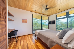 A modern bedroom with a wooden ceiling, a bed with a patterned bedspread, a desk with a chair, a TV mounted on the wall, and large windows offering a view of trees. - Exclusive 4-Villa Beach Estate 11 BR • Sleeps 22 (Pinilla)