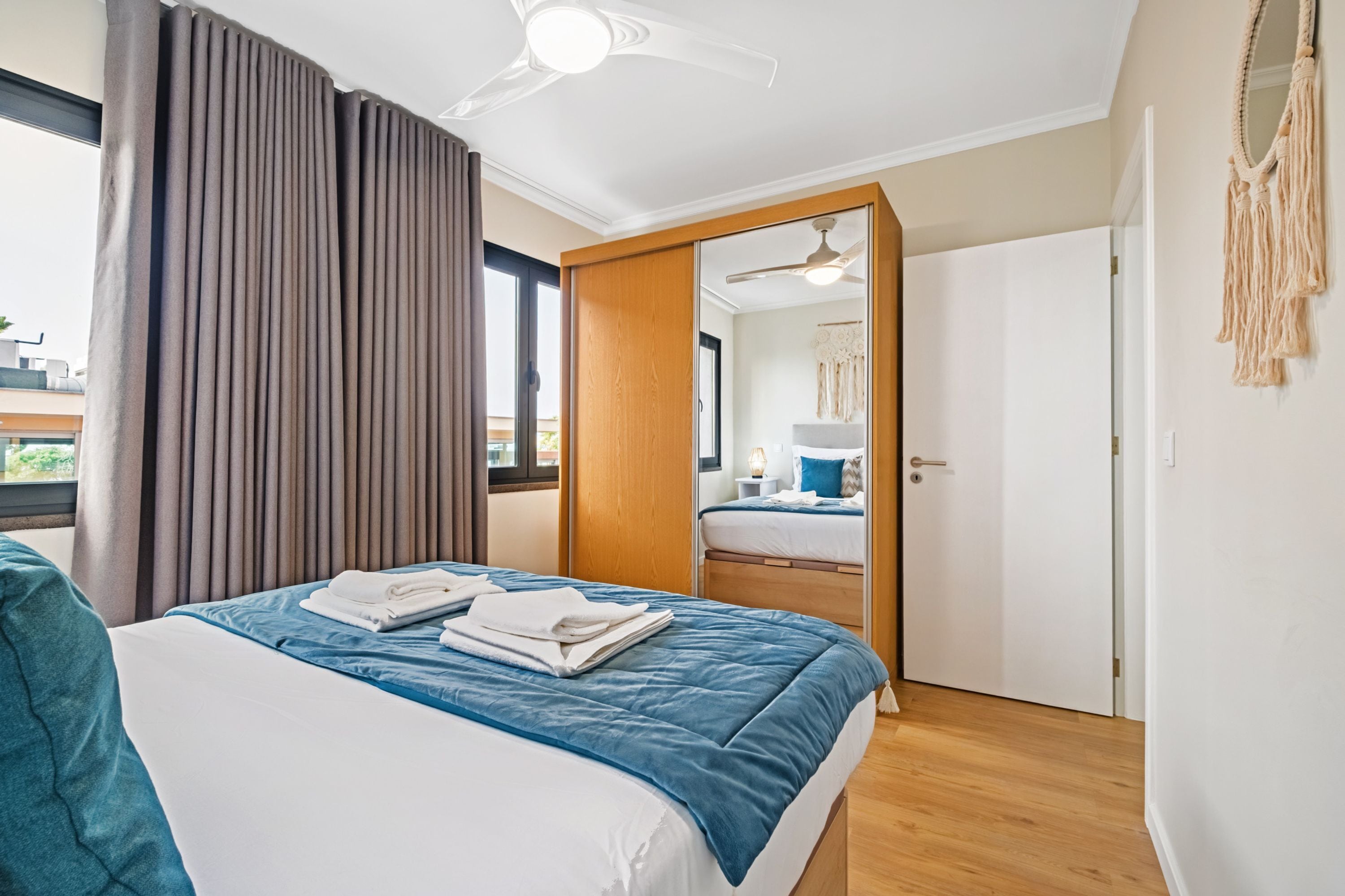 Quarto de hotel com cama, ventilador de teto, janela com cortinas e porta que dá para outro cômodo.