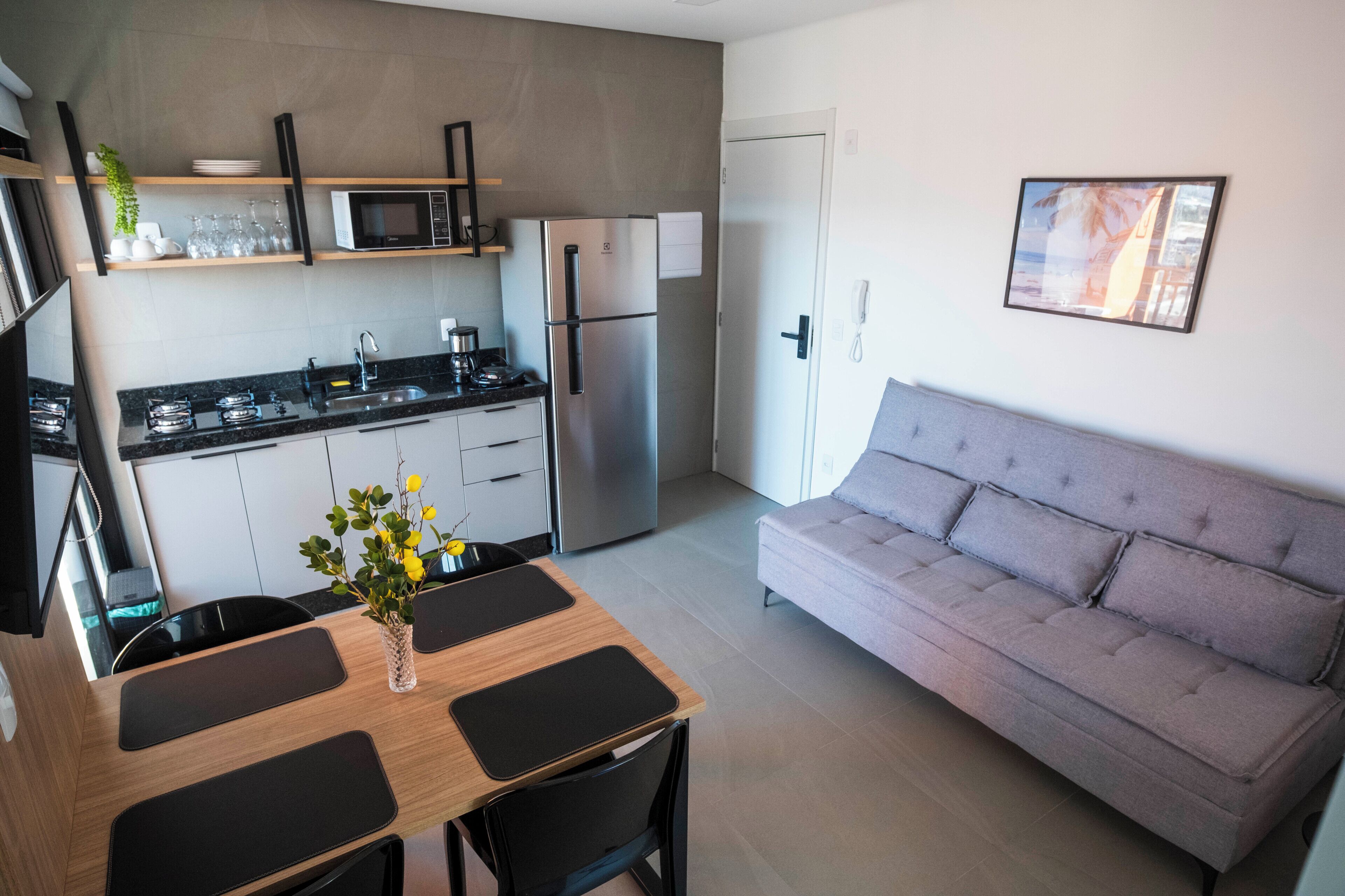 A modern kitchen with a refrigerator, microwave, and stove. There is a dining table with chairs and a vase of flowers. A gray sofa is against the wall.