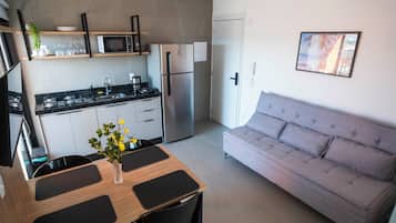 A modern kitchen with a refrigerator, microwave, and stove. There is a dining table with chairs and a vase of flowers. A gray sofa is against the wall.