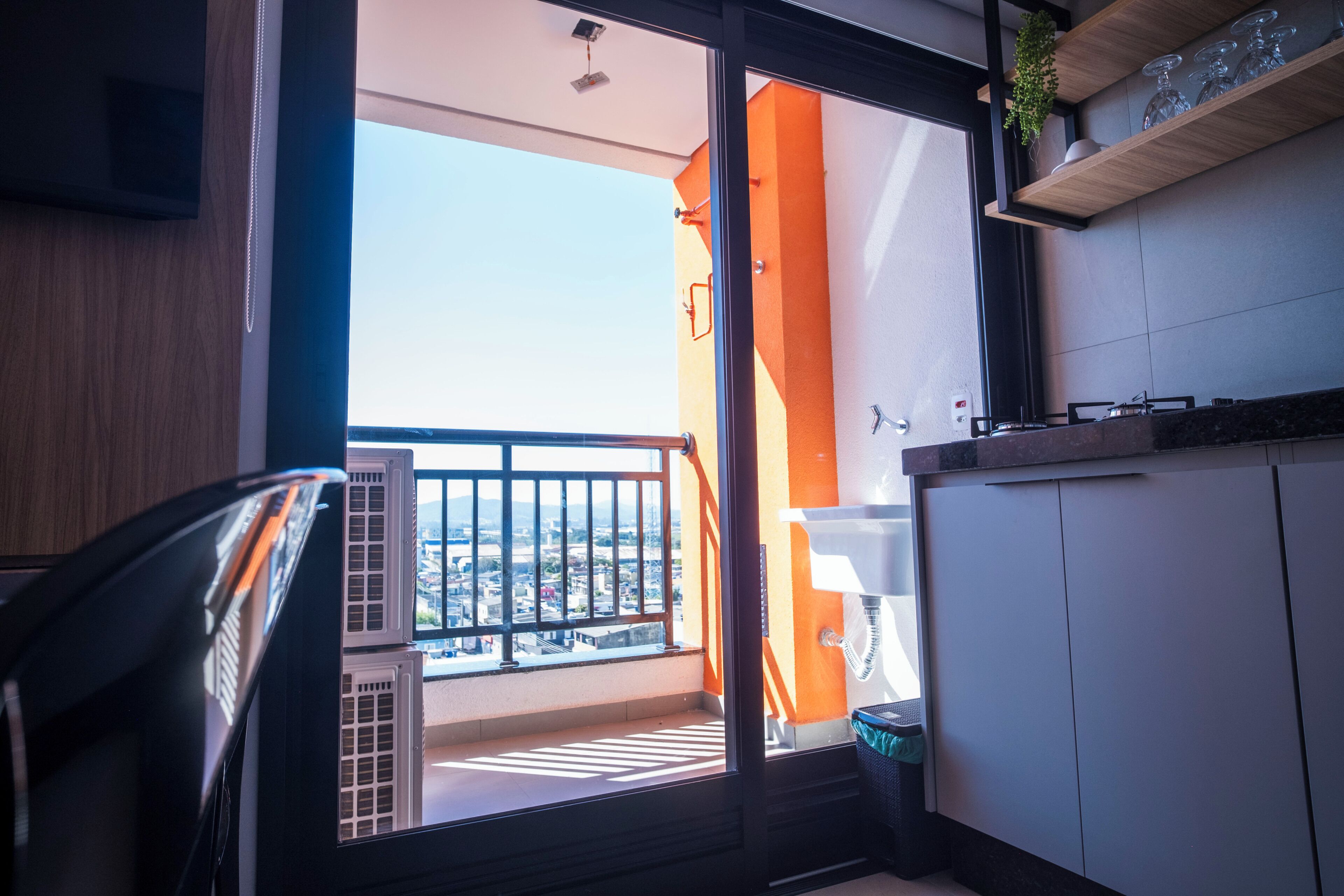A balcony with a view of the city, a window with blinds, and a small trash bin.