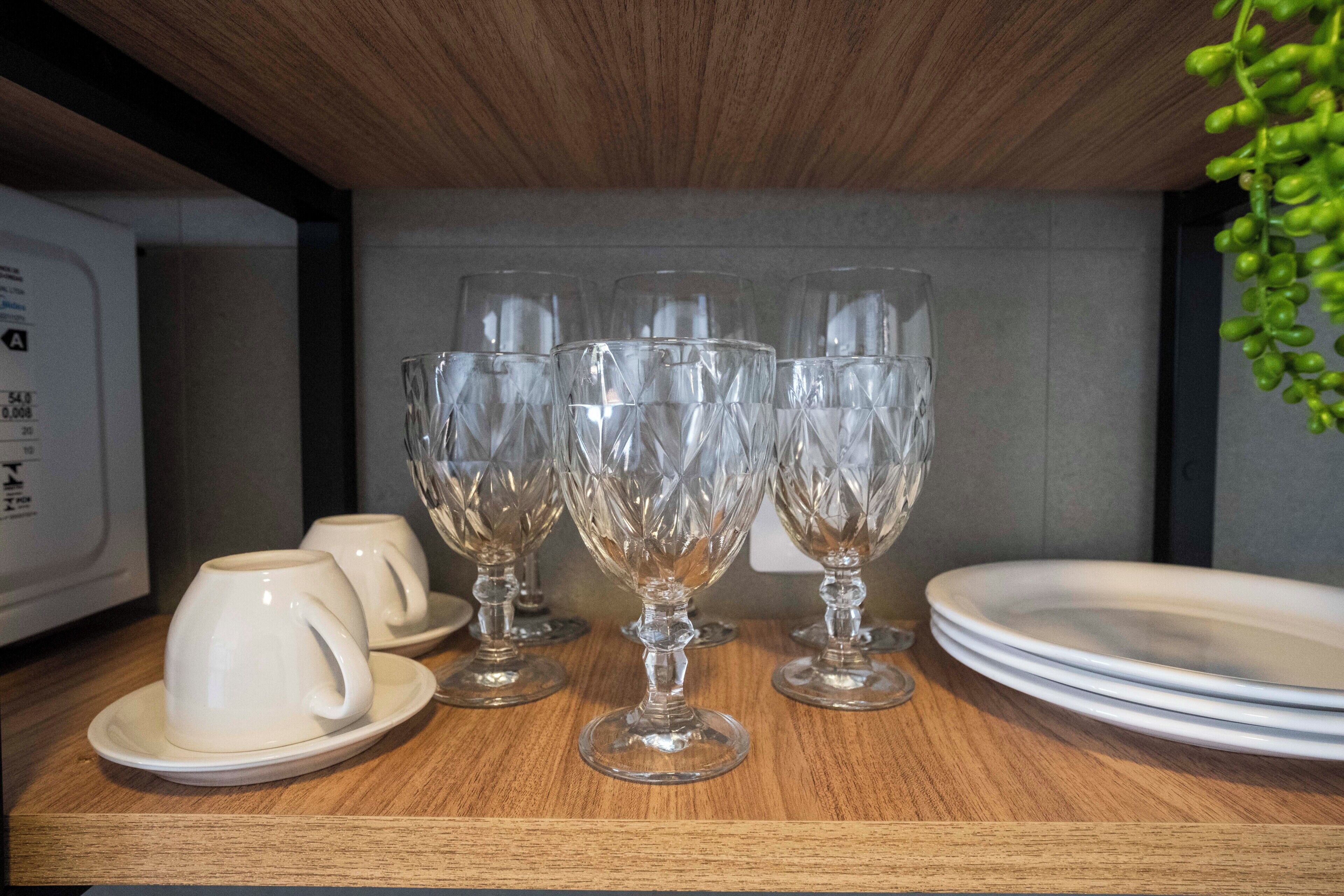 A wooden shelf with four clear glass goblets and two white ceramic cups with saucers.