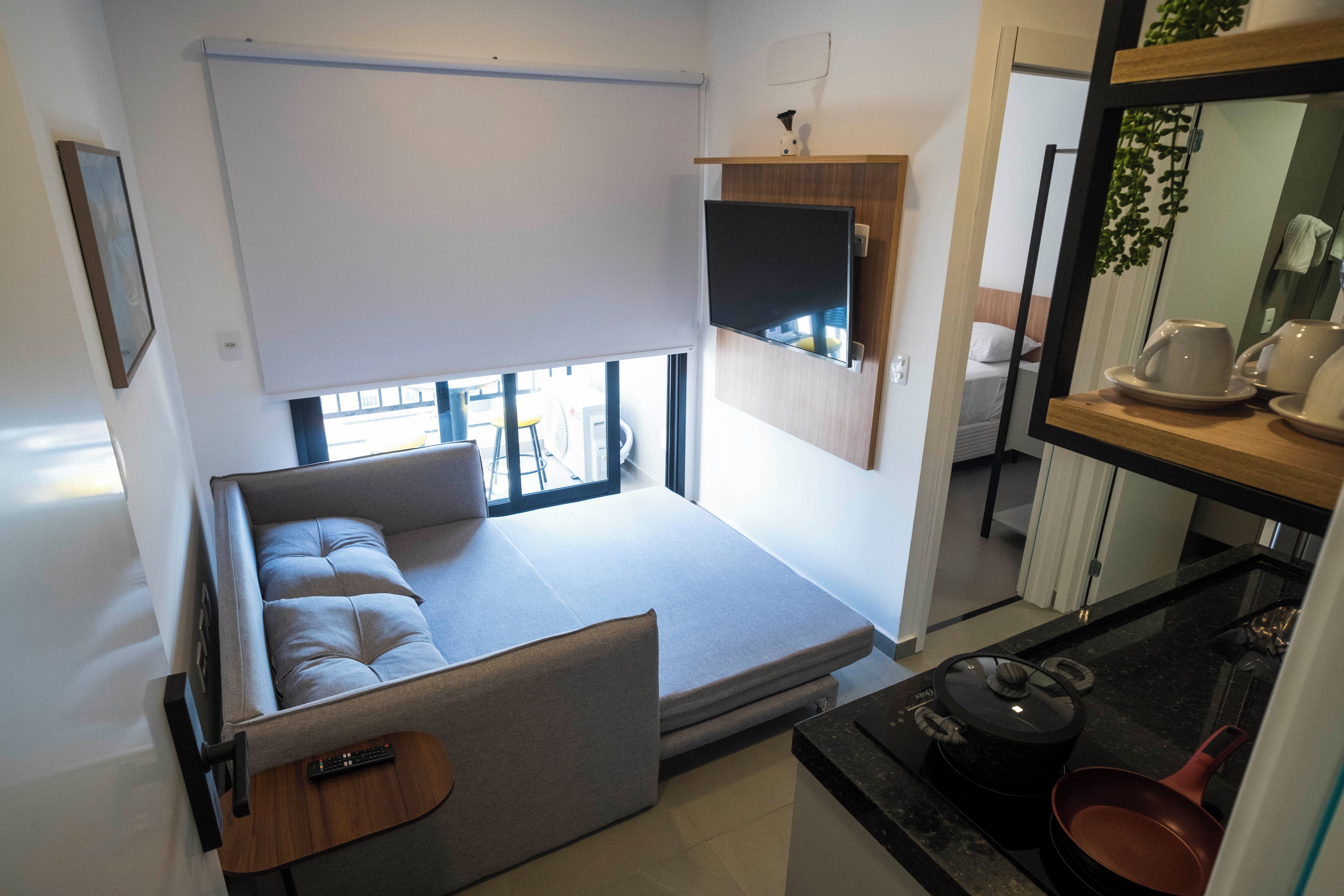 A compact living space with a sofa, a flat-screen TV mounted on the wall, a small kitchen area with a stove and pots, and a window providing natural light.