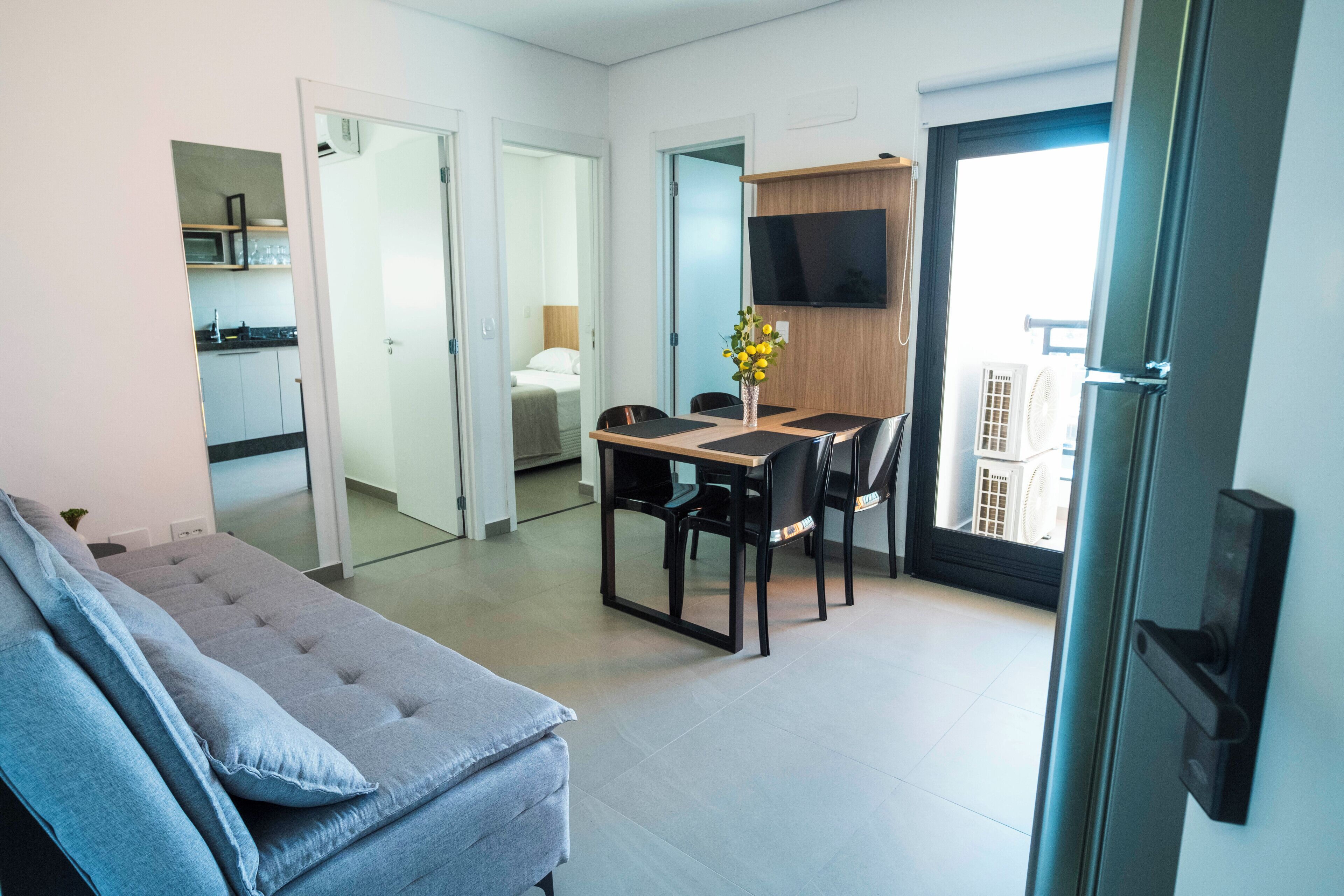 Una sala de estar moderna con un sofá gris, un comedor con una mesa y sillas de madera, un televisor montado en la pared y vistas de la ciudad a través de un ventanal grande.