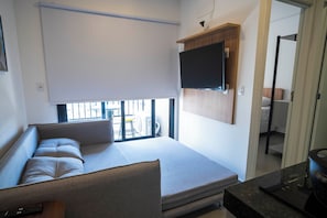 A modern living room with a sofa, a flat-screen TV mounted on the wall, and a window with blinds. - Vista Cecap (Guarulhos)