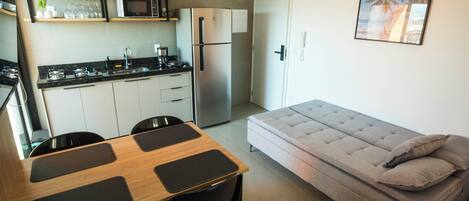 Un espacio habitable compacto con kitchenette, una mesa de comedor con cuatro sillas, un sofá con cojines y un cuadro en la pared.